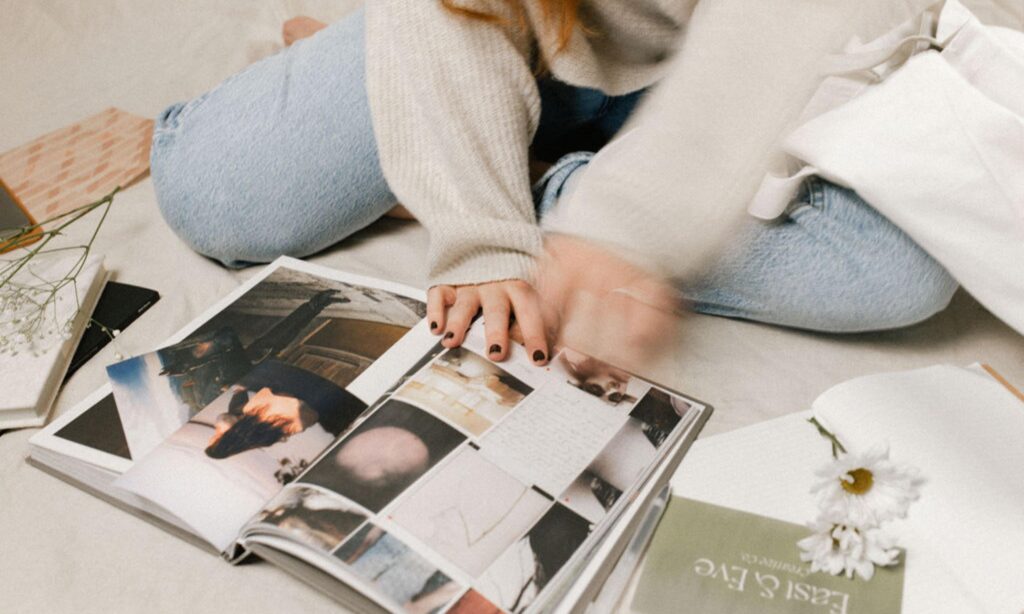 A photo of founder, Madeline - flipping through an art book on the floor with film photographs and cards with East & Eve Creative Co's branding on them nearby.